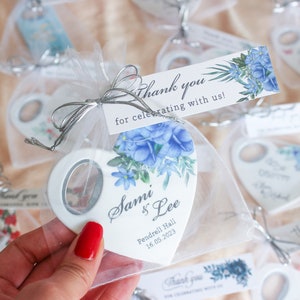 Könnte beinhalten: Weißer herzförmiger Flaschenöffner mit blauem Blumenmuster und dem Text "Sami & Lee" und "Pendrell Hall 16.05.2023". Der Flaschenöffner ist in einem weißen Organzasack mit silbernem Band verpackt.