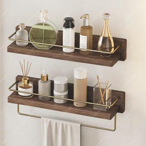 Puede incluir: Dos estantes de baño de madera oscura con detalles metálicos dorados. El estante superior contiene frascos de perfume y productos para el cuidado de la piel. El estante inferior tiene un toallero con una toalla blanca doblada y más artículos de tocador.