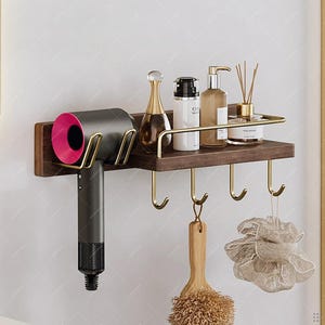 May include: A dark brown wooden wall-mounted shelf with gold-colored metal accents. It features a hairdryer holder, a shelf for toiletries, and hooks for hanging items. A hairdryer with a pink accent is in the holder.