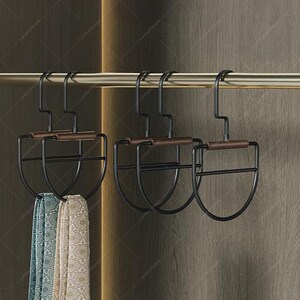 May include: Black metal scarf hangers with brown wooden accents are suspended from a gold-colored rod. Two patterned scarves in shades of blue, green, and beige hang from the hangers. The hangers have a unique, curved design.