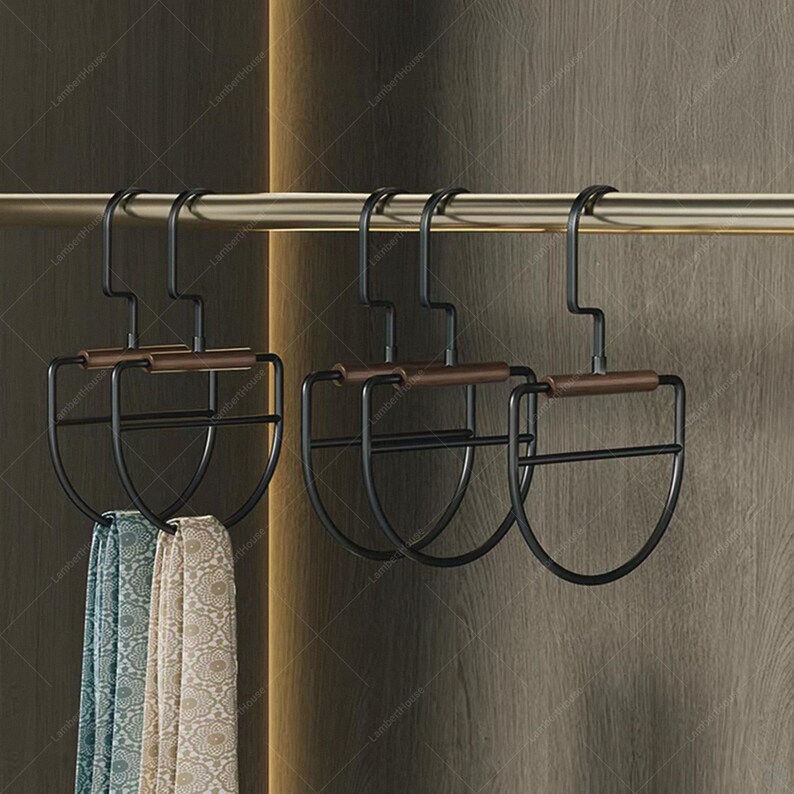 May include: Black metal scarf hangers with brown wooden accents are suspended from a gold-colored rod. Two patterned scarves in shades of blue, green, and beige hang from the hangers. The hangers have a unique, curved design.