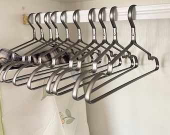 May include: A set of 12 silver metal hangers with a smooth, rounded design. The hangers are stacked on top of each other and are hanging from a white shelf.