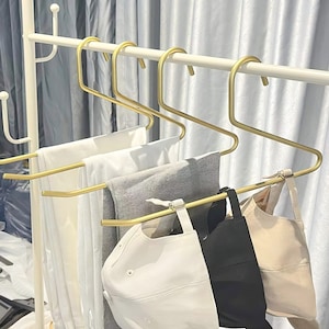 Op de afbeelding: Gouden metalen hangers met een uniek S-vormig ontwerp, die een verscheidenheid aan kledingstukken vasthouden, waaronder witte T-shirts, grijze broeken en een witte en zwarte baseballpet.