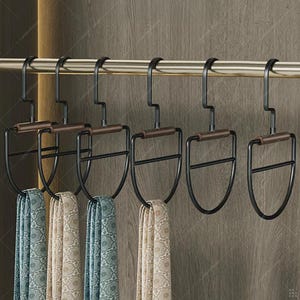 May include: A set of six black metal scarf hangers with brown wooden handles, hanging on a gold-colored rod. Each hanger holds a patterned scarf in shades of blue and beige. The hangers are designed to organize scarves.