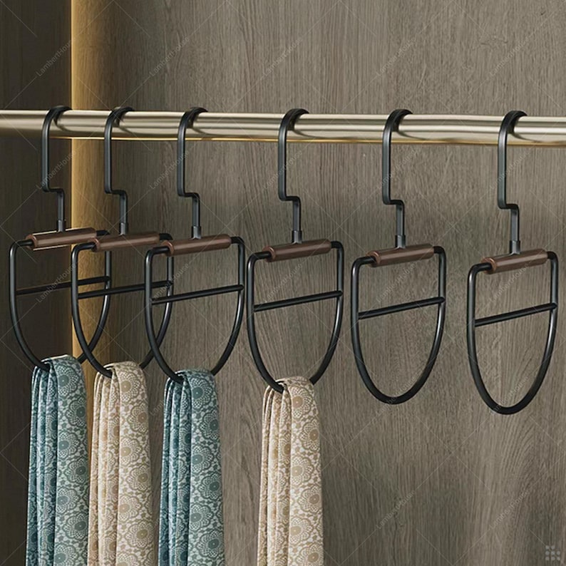 May include: A set of six black metal scarf hangers with brown wooden handles, hanging on a gold-colored rod. Each hanger holds a patterned scarf in shades of blue and beige. The hangers are designed to organize scarves.