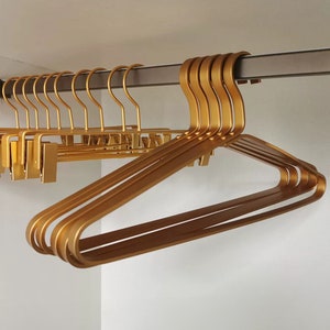May include: A set of gold metal hangers with a slim, modern design. The hangers are stacked on top of each other and are hanging from a metal rod.