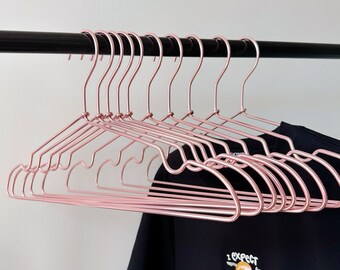 May include: A set of 10 rose gold metal hangers. The hangers are thin and have a rounded shape. They are hanging on a black metal rod.