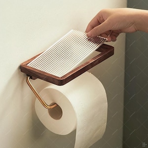 May include: A wooden and glass toilet paper holder with a roll of white toilet paper. The holder has a rectangular frame with a dark wood finish and a ribbed glass top. The metal arm is gold-colored.