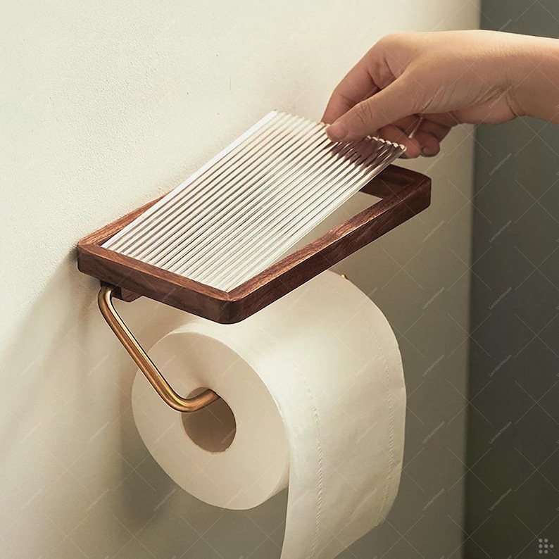 May include: A wooden and glass toilet paper holder with a roll of white toilet paper. The holder has a rectangular frame with a dark wood finish and a ribbed glass top. The metal arm is gold-colored.