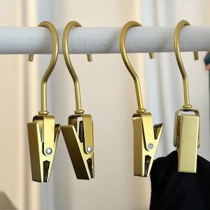 May include: Four gold-colored metal clips with hooks hanging from a white bar. The clips have a spring-loaded mechanism to hold items.
