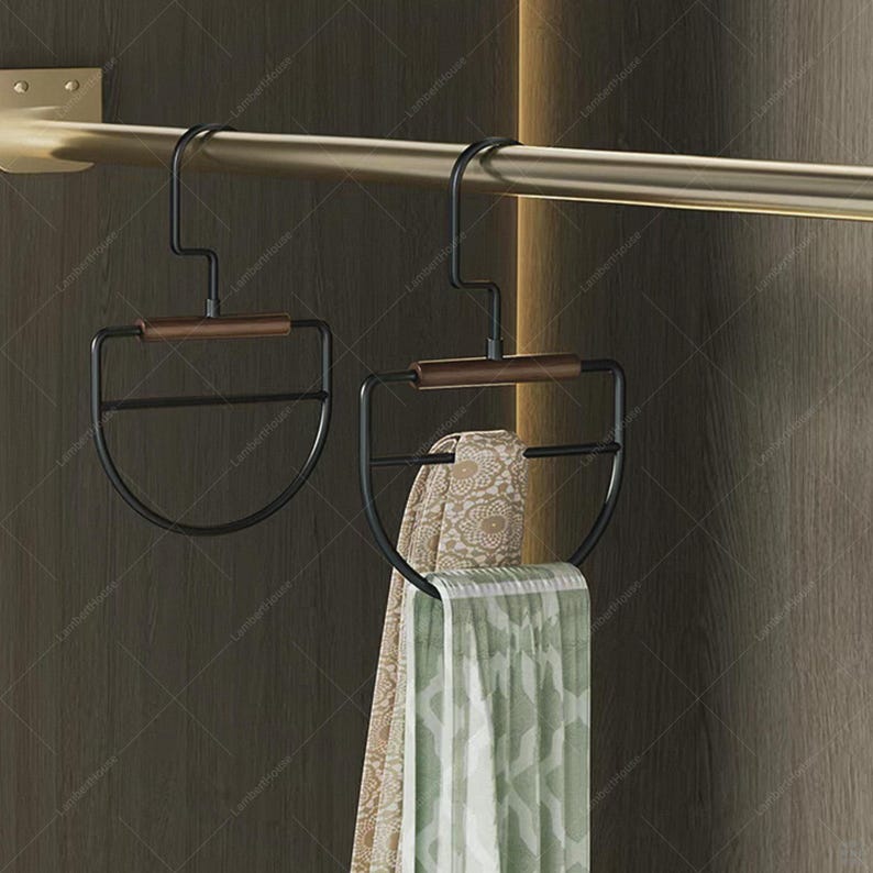 May include: Two black metal scarf hangers with wooden accents are suspended from a gold-colored rod. One hanger holds a patterned scarf, and the other is empty. The hangers have a semi-circular design.