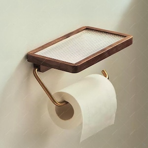 May include: A wall-mounted toilet paper holder with a rectangular wooden shelf and a brass-colored metal arm. The shelf has a textured glass insert. A roll of white toilet paper is in place.