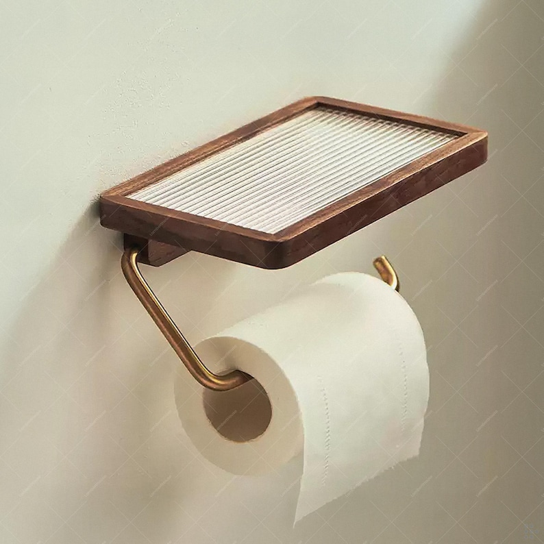 May include: A wall-mounted toilet paper holder with a rectangular wooden shelf and a brass-colored metal arm. The shelf has a textured glass insert. A roll of white toilet paper is in place.