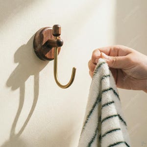 May include: A wooden wall hook with a dark brown finish and a brass hook. The hook is mounted on a light-colored wall. A hand is holding a white towel with green stripes, ready to hang it on the hook.