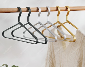 May include: A set of six metal clothes hangers in black, silver, and gold finishes, each featuring a smooth, angular frame and a curved hook. The hangers are suspended from a wooden rod, with a white textured sweater hanging on the right side.