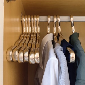 Könnte beinhalten: Nahaufnahme eines Kleiderschranks mit mehreren goldenen Kleiderbügeln, an denen verschiedene Kleidungsstücke hängen, darunter ein weißes Hemd mit Knöpfen, ein hellblaues Hemd, ein dunkelblaues Hemd und ein grünes Hemd.