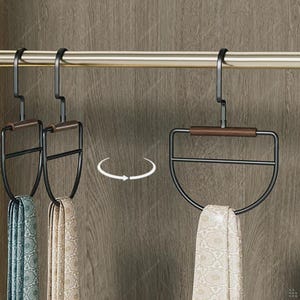 May include: Black metal tie hangers with a wooden accent, holding patterned scarves. The hangers are suspended from a gold-colored rod, with a wooden cabinet in the background. The design is modern and functional.