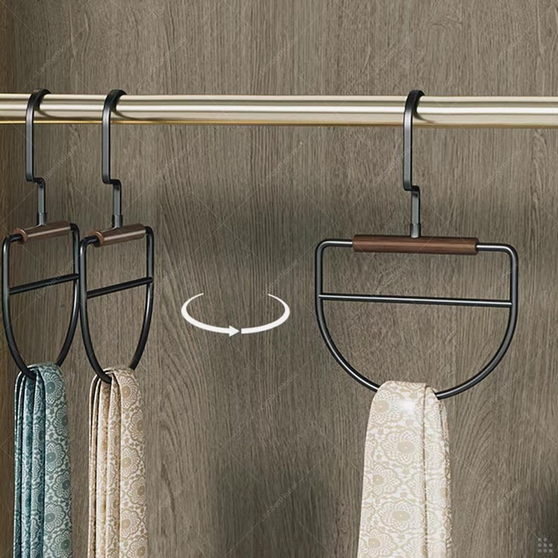 May include: Black metal tie hangers with a wooden accent, holding patterned scarves. The hangers are suspended from a gold-colored rod, with a wooden cabinet in the background. The design is modern and functional.