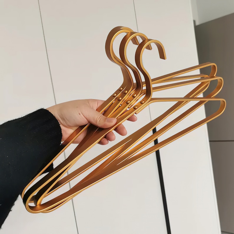 May include: Four gold metal hangers with a rounded shape and a textured surface. The hangers are stacked on top of each other.