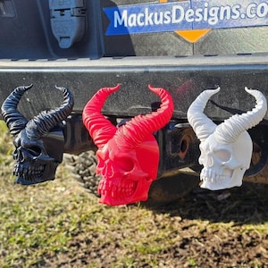 Puede incluir: Tres cubiertas de enganche de remolque con forma de calavera en negro, rojo y blanco. Cada calavera tiene cuernos grandes y curvos. Las cubiertas de enganche están montadas en un enganche de vehículo negro. El sitio web MackusDesigns.com es visible en el fondo.