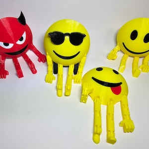 Op de afbeelding: Vier 3D-geprinte figuren, drie zijn gele smiley-gezichten met verschillende uitdrukkingen, één is rood met hoorns en boze ogen. Elke figuur heeft scharnierende armen en benen.
