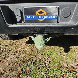 Puede incluir: Una tapa de enganche de cabeza de Yoda verde unida a un enganche de camión negro. El camión tiene una placa negra con el texto "MackusDesigns.com" en ella.