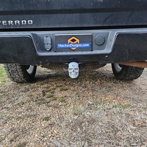 May include: A black truck's rear with a chrome emblem and the word "Silverado". A skull-shaped trailer hitch cover is visible below the bumper. A rectangular plate with the text "MackusDesigns.com" is also visible.