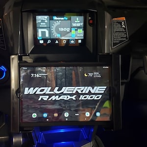 Puede incluir: Un tablero negro con una pantalla digital que muestra una velocidad de 1300 y un nivel de batería del 168%. Una tableta está montada en el tablero y muestra el texto "WOLVERINE RMAX 1000".