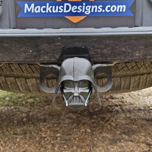 Puede incluir: Una tapa de enganche de cabeza de Darth Vader de plástico negro unida a un parachoques de coche negro. La tapa de enganche es un accesorio popular para los entusiastas de los coches.