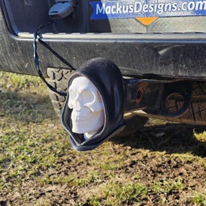 Pode incluir: Uma caveira branca com capuz preto, presa a um engate de reboque. A caveira é impressa em 3D. O engate está montado num pára-choques preto. O fundo mostra relva e o site "MackusDesigns.com".