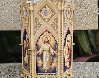Puede incluir: Una linterna decorativa beige con forma de catedral, cortada con láser. La linterna presenta paneles de estilo vidriera que representan figuras religiosas. La parte superior tiene una cruz y cruces más pequeñas. La linterna está hecha de madera clara y se encuentra sobre una superficie de piedra.