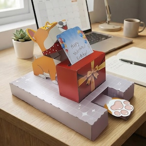 May include: A papercraft corgi dog holding a card that reads "Happy Valentine/Birthday!" The dog is positioned above a red gift box with a gold bow. The box sits on a light purple platform with a paw-shaped pull-out. A laptop and desk lamp are in the background.