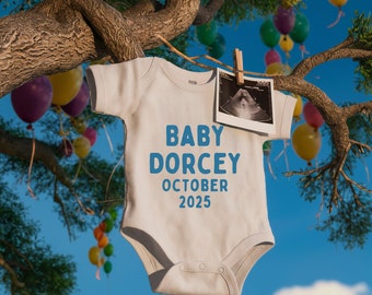 May include: A white baby onesie with the text "BABY DORCEY OCTOBER 2025" printed in blue. A black and white ultrasound image is hanging from a clothesline with a clothespin.
