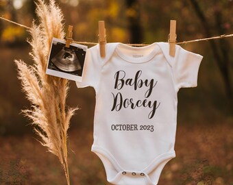 Könnte beinhalten: Weißer Baby-Strampler mit schwarzem Text, der "Baby Dorcey October 2023" lautet. Ein Schwarzweiß-Ultraschallbild hängt neben dem Strampler.