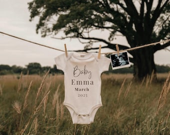 May include: A white baby onesie with the text "Baby Emma March 2025" hanging on a clothesline in a field of tall grass. A black and white ultrasound image is hanging on the clothesline next to the onesie.