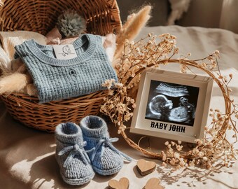 May include: A wicker basket filled with a blue knitted sweater, a framed ultrasound image with the text "Baby John", and two pairs of blue knitted baby booties. The basket is surrounded by dried flowers.