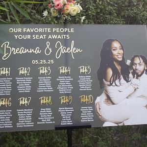Könnte beinhalten: Ein Hochzeits-Sitzplan mit den Namen Breanna & Jaelen, datiert auf den 05.25.25. Das Schild hat goldfarbene Schrift und enthält ein Foto des Paares. Die Tabelle ist in zehn Tische unterteilt, jeder mit einer Liste von Namen.
