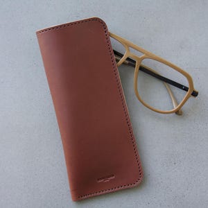 May include: A brown leather glasses case with a stitched edge and a small logo. The case is lying on a light gray surface with a pair of light brown glasses next to it.