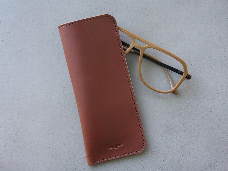 May include: A brown leather glasses case with a stitched edge and a small logo. The case is lying on a light gray surface with a pair of light brown glasses next to it.