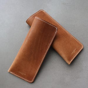 May include: Two rectangular tan leather cases with rounded corners and white contrast stitching along the edges. Each case features a small embossed logo near the bottom, showcasing a smooth finish and a slim, elongated design for storage.