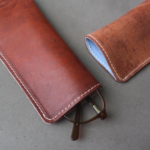 May include: A brown leather glasses case with white stitching, holding a pair of eyeglasses. The case is open, revealing the glasses. Another case is visible, with a light blue interior.