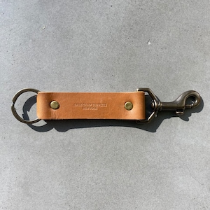May include: A brown leather key chain with a brass key ring and a brass hook. The key chain has the text "BASE CAMP SUPPLIES NEW YORK" embossed on it.