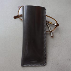 May include: A dark grey leather glasses case with a pair of spectacles resting on top. The case is rectangular with a stitched border. The spectacles have brown frames and clear lenses.