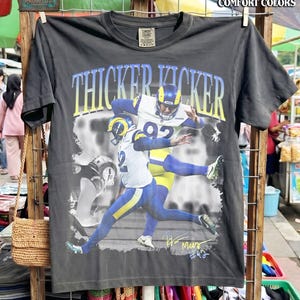 May include: A charcoal gray short-sleeve t-shirt featuring a graphic of a football player in a white, blue, and yellow uniform. The text THICKER KICKER is printed in blue and yellow above the player, with a signature and the number 92 below.