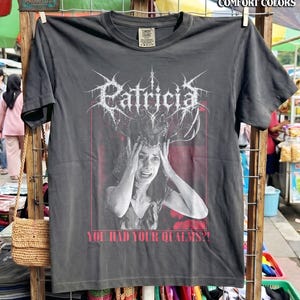 May include: A charcoal gray short-sleeve t-shirt featuring a black and white graphic of a person holding their head with both hands, topped with deer antlers. The text "Patricia" in a stylized font and "YOU HAD YOUR QUALMS?!" are printed.