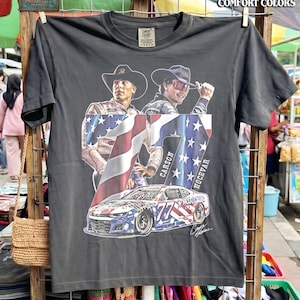 May include: Dark gray t-shirt featuring a graphic of two men in cowboy hats, an American flag, and a race car with the number 77. The shirt has the text "PEPPER COMFORT COLORS" and "PROJECT X CREATIVE".