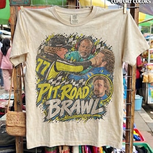 May include: A cream-colored t-shirt featuring a graphic print of a race car pit road brawl. The design includes three individuals, a checkered flag pattern, and the text Pit Road Brawl. The shirt has a crew neck and short sleeves.