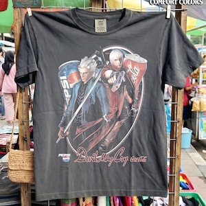Peut inclure: T-shirt gris foncé avec un motif de deux personnages de Devil May Cry, des canettes de Pepsi et de Coca-Cola. Le t-shirt porte l'inscription "Devil May Cry" et "Coca-Cola" dans un cercle. Le t-shirt est accroché sur un présentoir en bois.