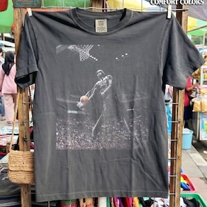 May include: A charcoal gray short-sleeve t-shirt featuring a graphic print of a dark armored figure dunking a basketball into a hoop in a crowded arena. The shirt has a crew neckline and a Comfort Colors brand label on the inside of the collar.