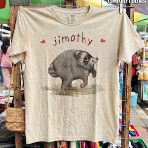 Pode incluir: Uma t-shirt de manga curta na cor creme com um gráfico de uma criatura redonda, cinzenta e peluda a caminhar. O texto "jimothy" está impresso em castanho acima da criatura, ladeado por dois pequenos corações vermelhos.